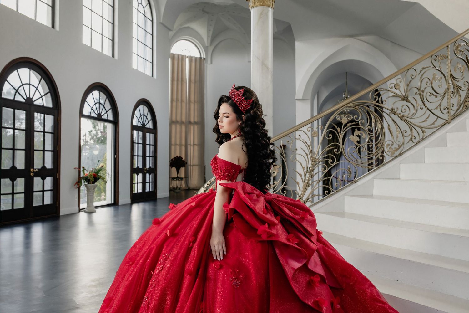 Elegant woman in a voluminous red quinceañera gown posing gracefully on a grand staircase in a luxurious white ballroom.