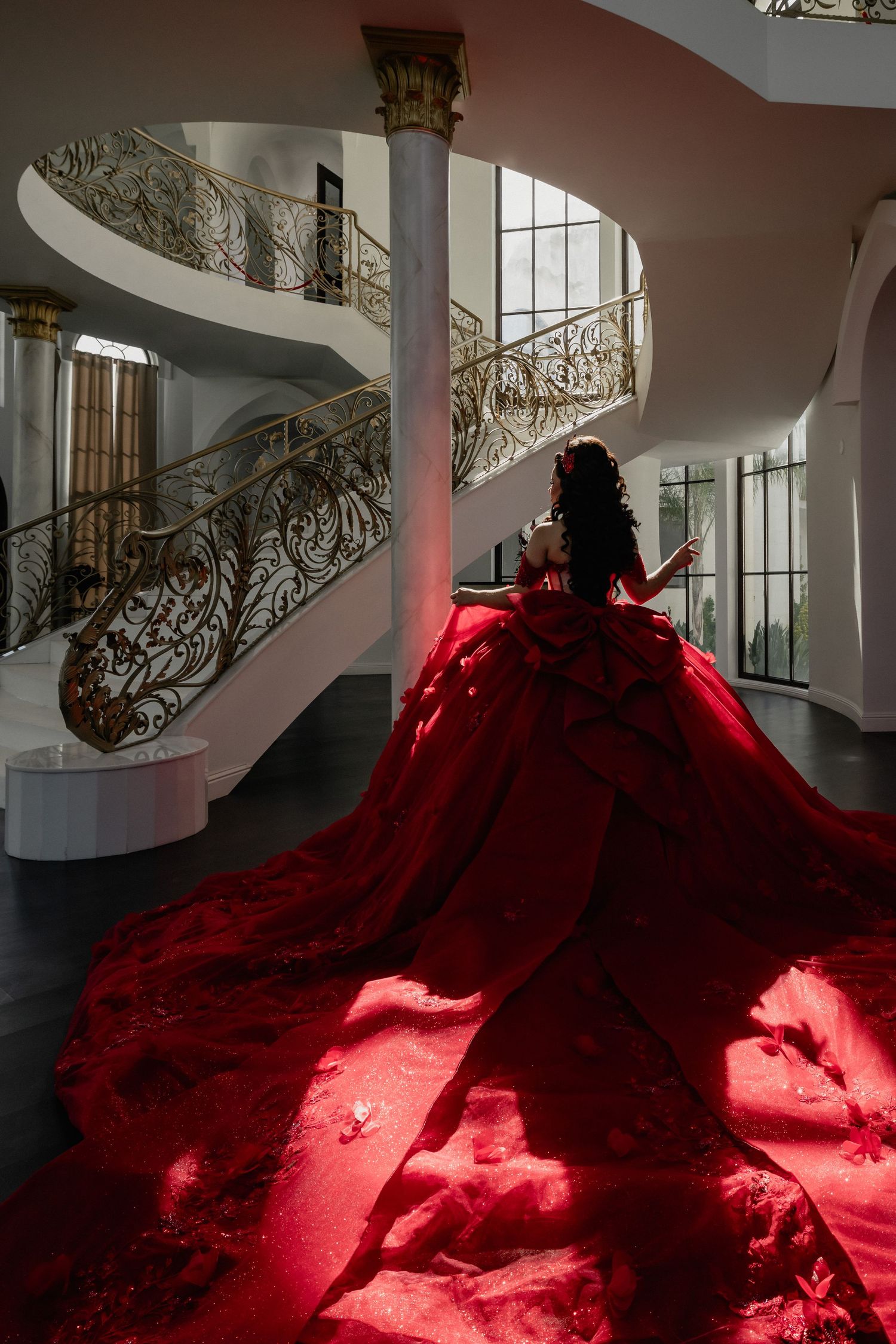 Young woman in a dramatic red ballgown with long train poses on grand staircase in luxurious white mansion.