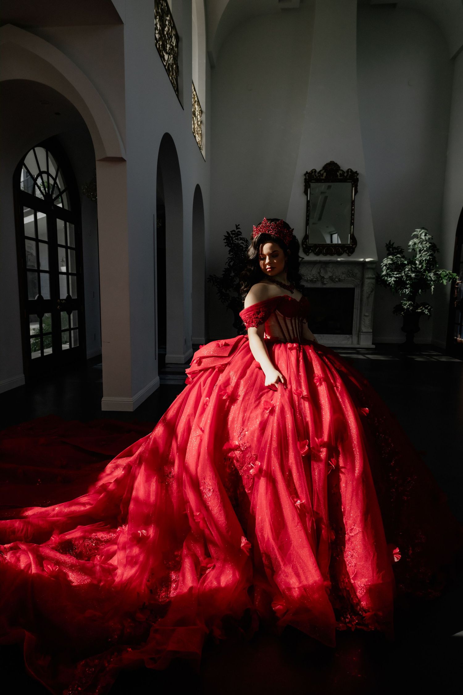 Woman wearing red ballgown and crown poses elegantly inside white castle interior.