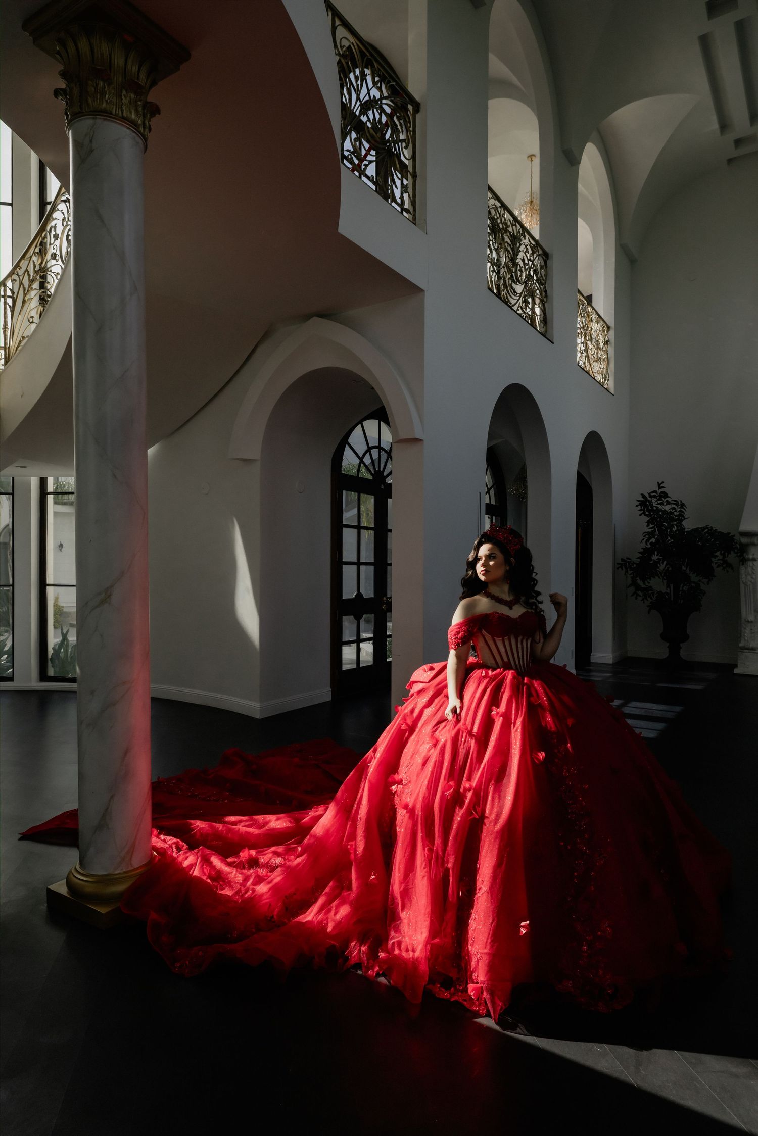 Woman in red ball gown with long train poses in elegant white marble hall with arched columns.