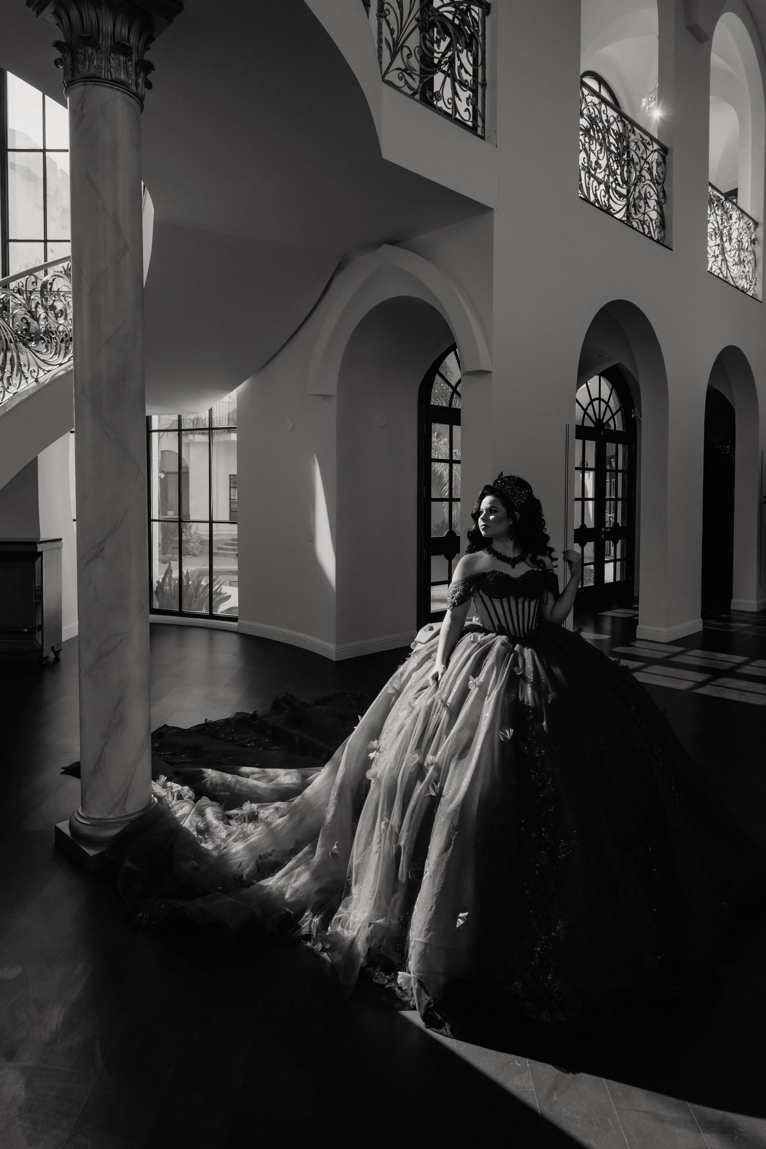 Elegant woman in voluminous ball gown stands in dramatic black and white interior with marble columns.