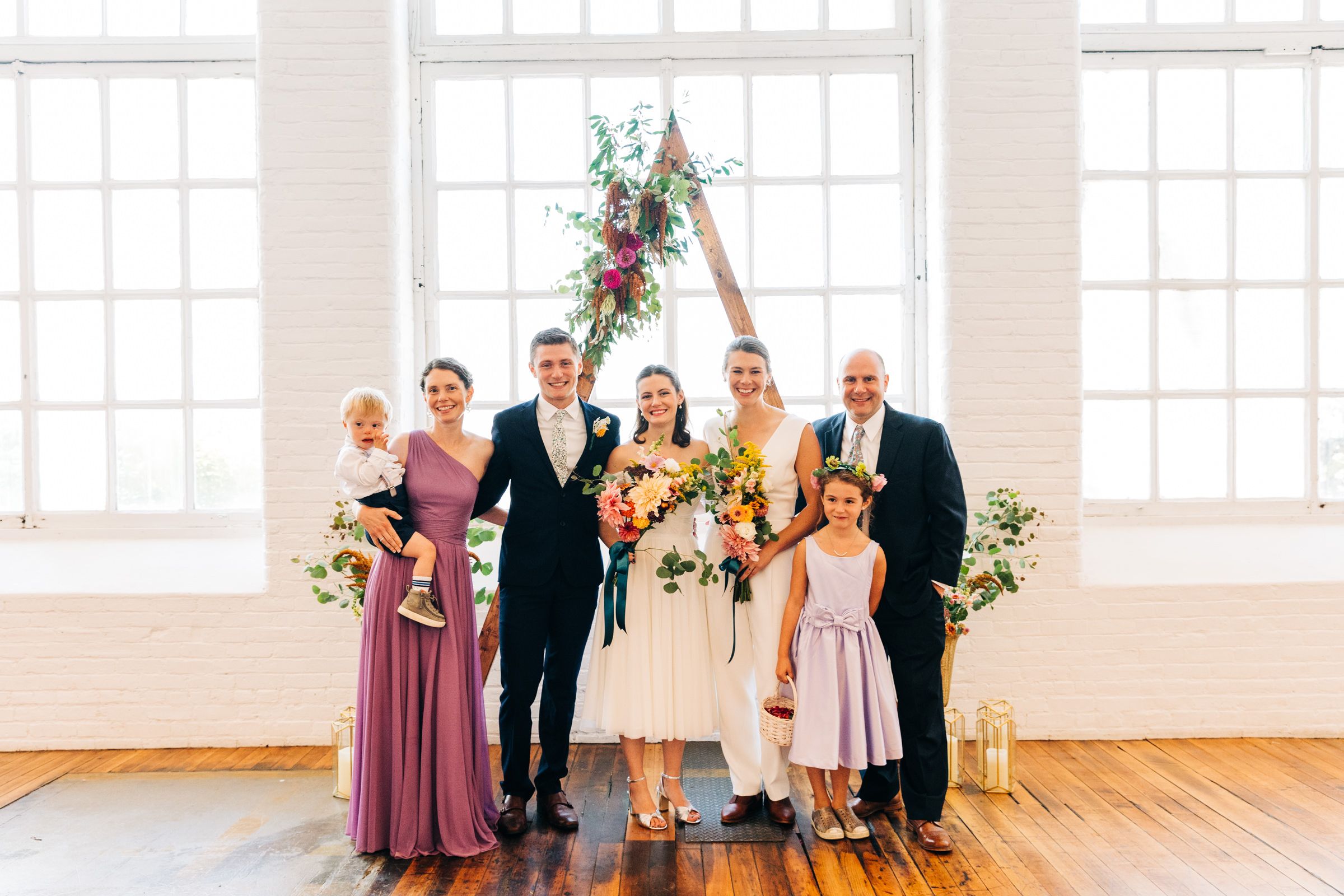 Wedding party lined up in formal attire with bridesmaids in mauve dresses against white brick wall.