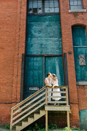 Couple sharing intimate moment on wooden stairs against rustic brick building with teal doors.