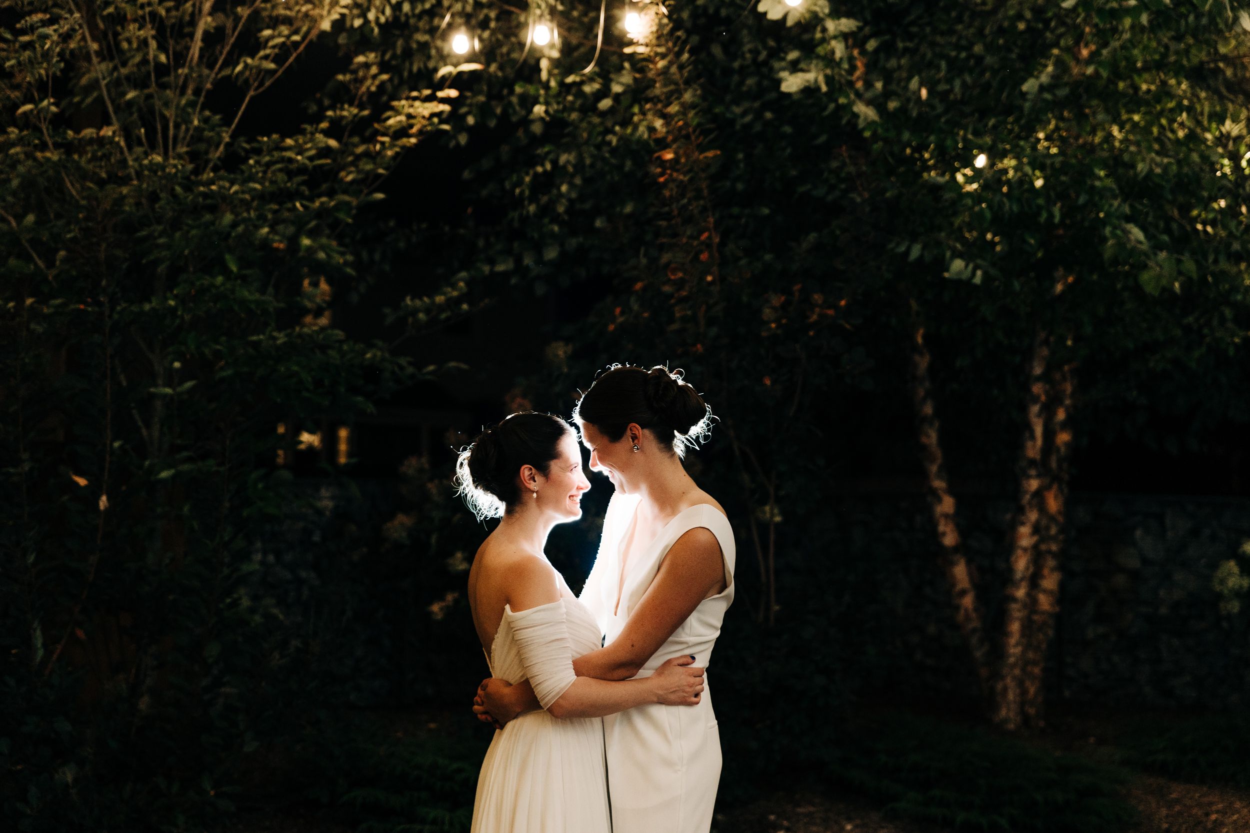 Two people in white dresses share an intimate moment at sunset among trees.