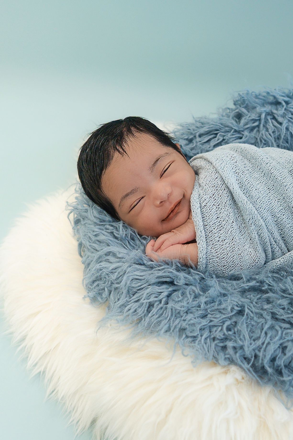 Smiling newborn baby swaddled in a light blue wrap, resting on a fluffy blue and white fur blanket.