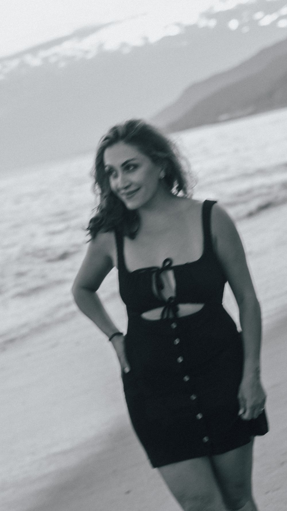Woman in black mini dress smiling on beach with snowy mountains in background, black and white photo.