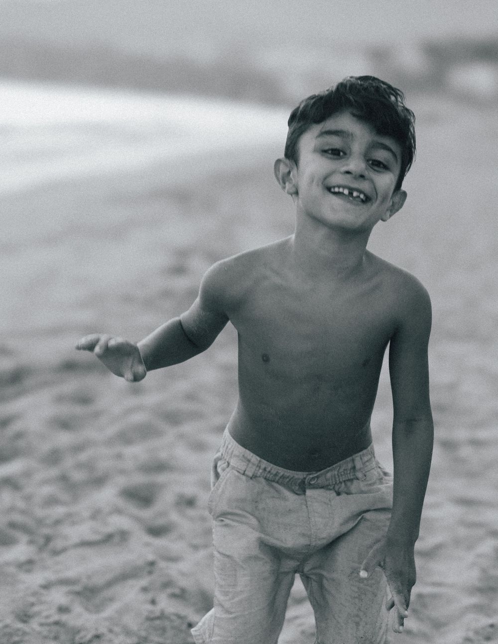 A smiling shirtless boy plays on a sandy beach in a vintage black and white photo.