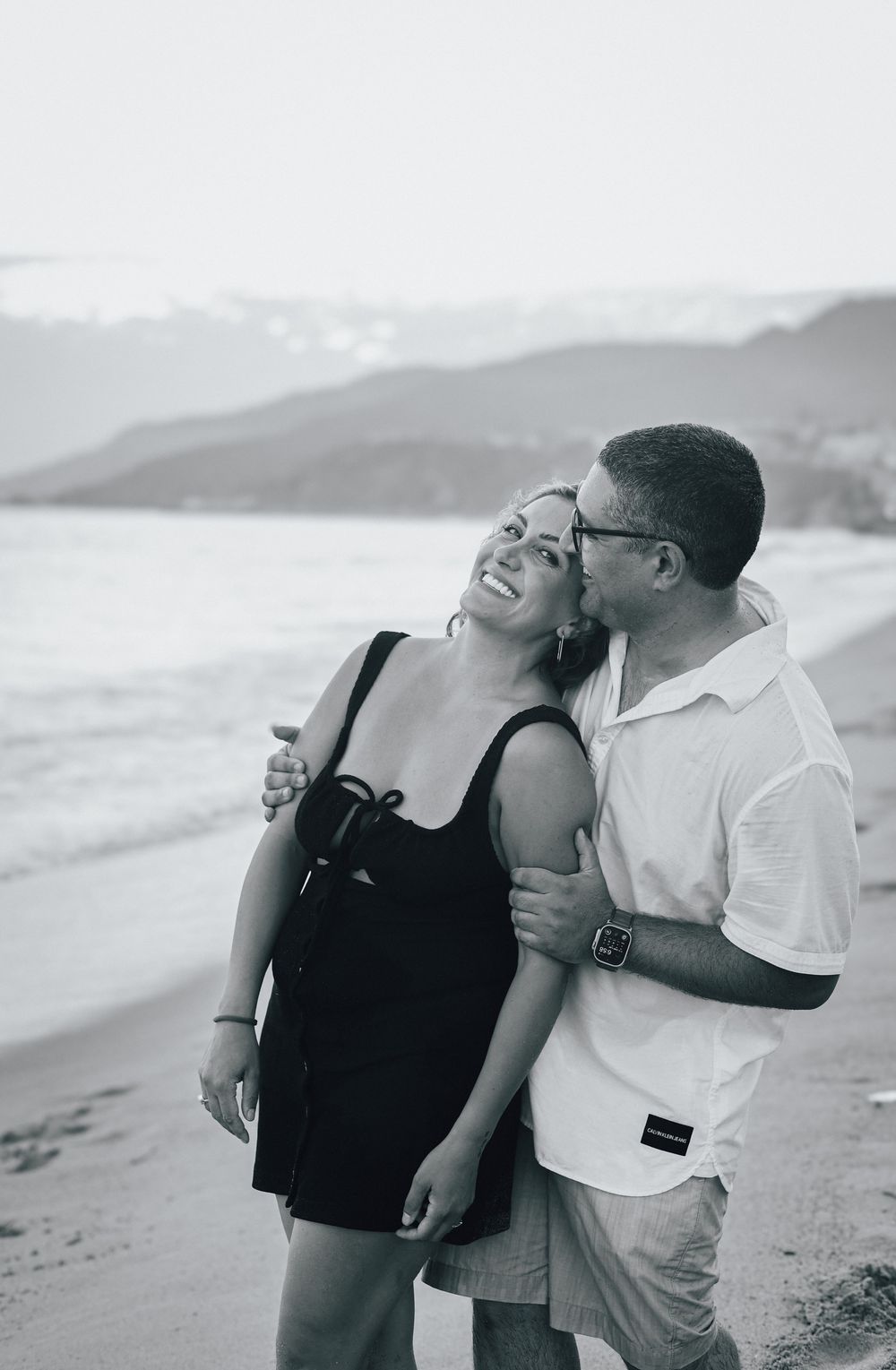 Black and white photo of a couple embracing and laughing on a beach with mountains in the background.