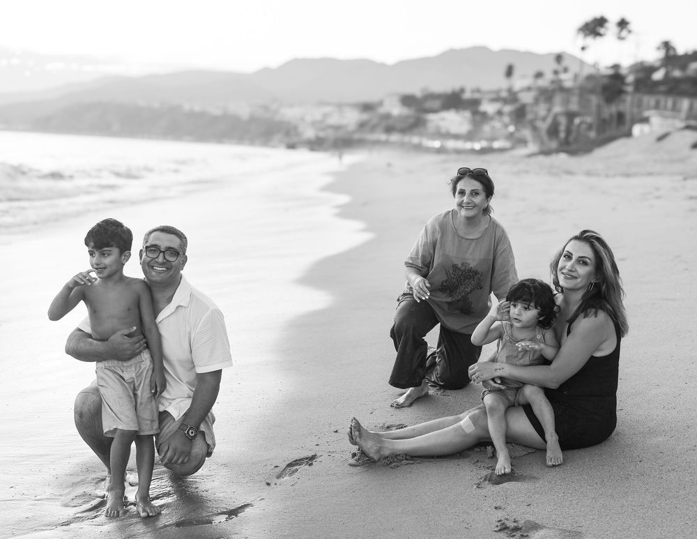 Black and white family beach photo with parents, grandmother, and two young children smiling on sandy shore.