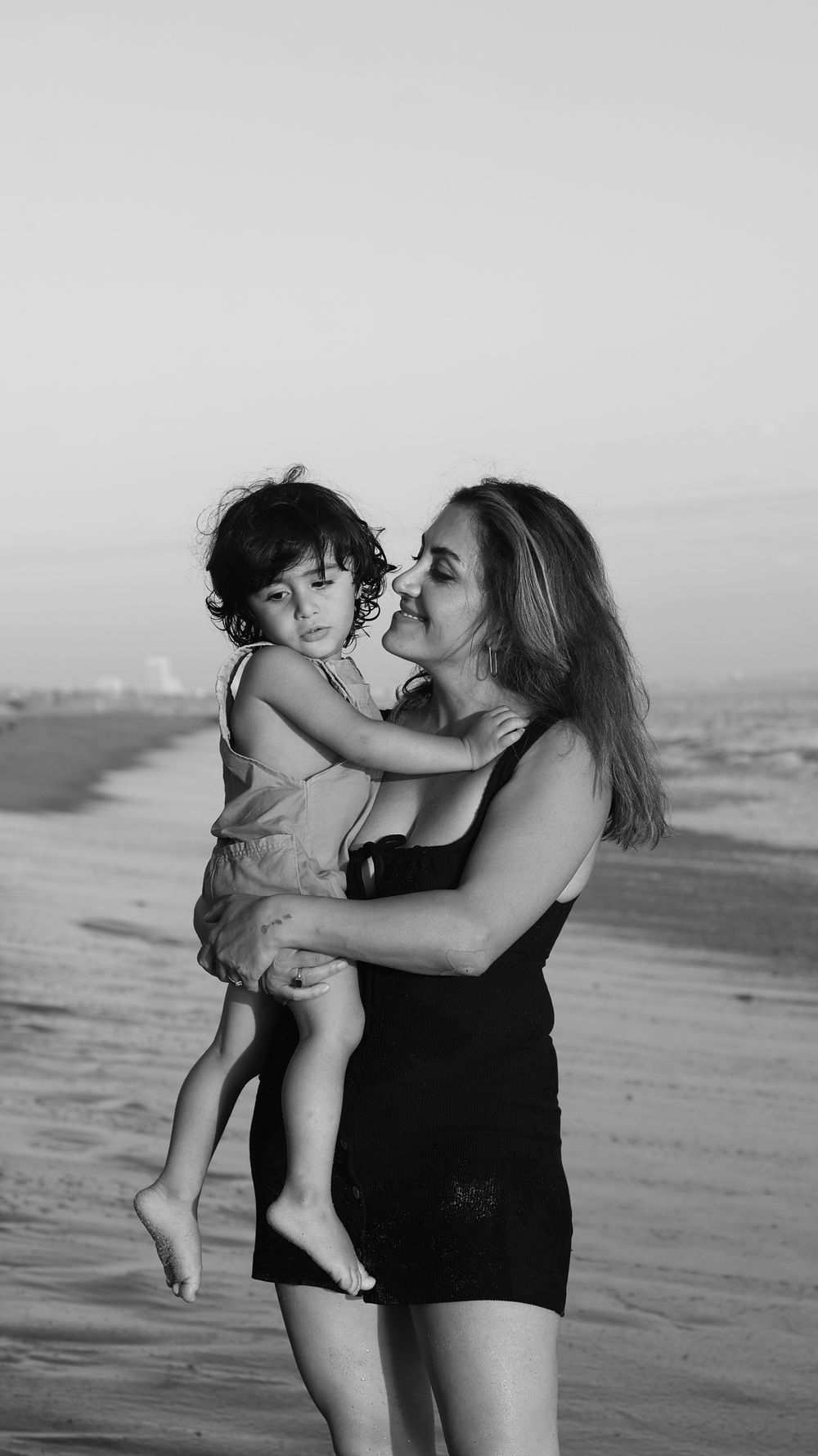 Woman in black dress holds young child on beach in black and white photo.