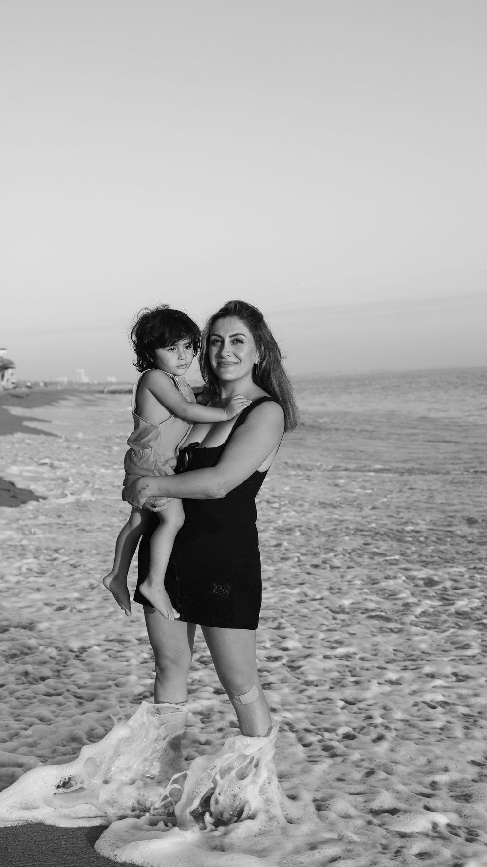 Black and white photo of a smiling woman holding a young child on a sandy beach with ocean waves.