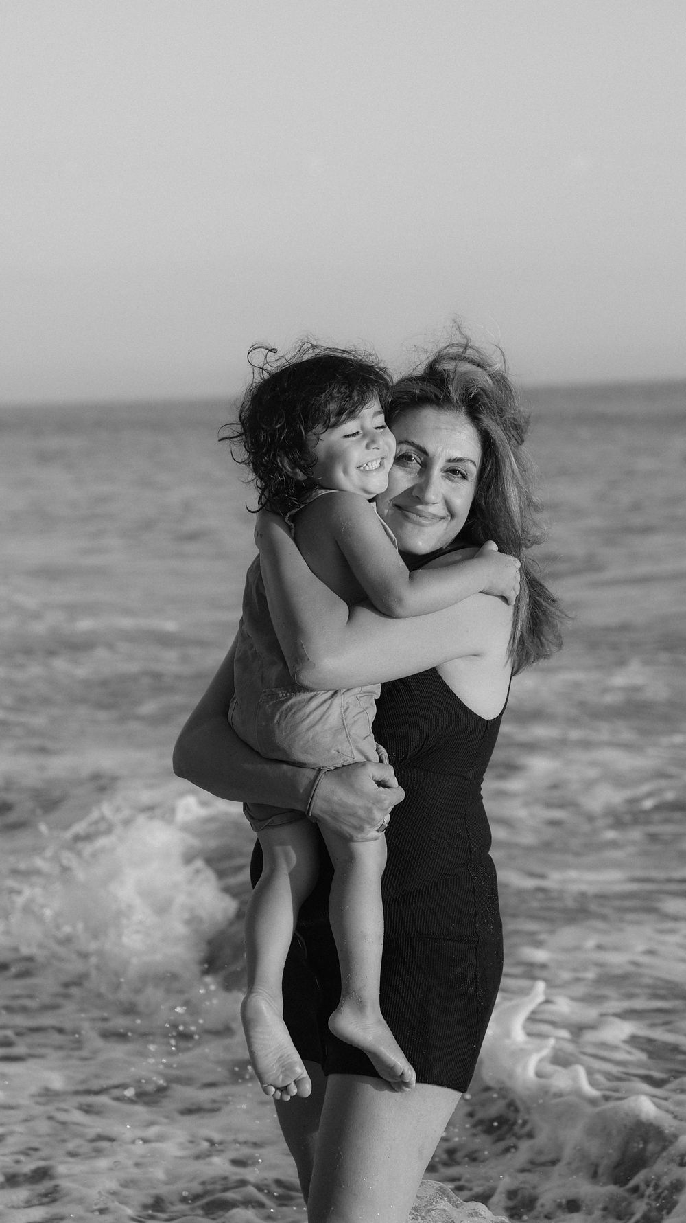A smiling mother holds her happy child on a beach in a black and white photo.