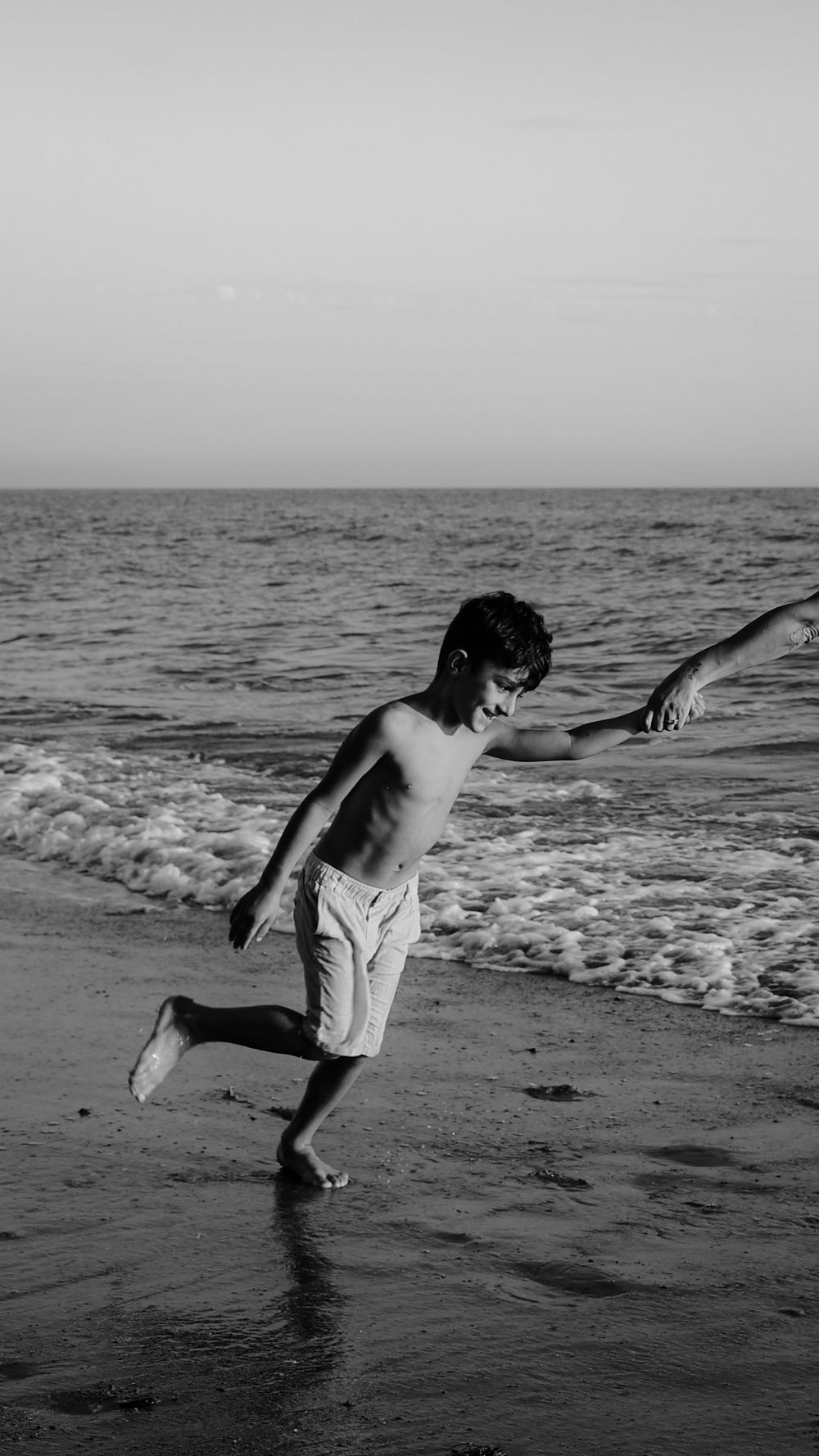 A shirtless boy plays joyfully in ocean waves on a sandy beach, captured in black and white photography.