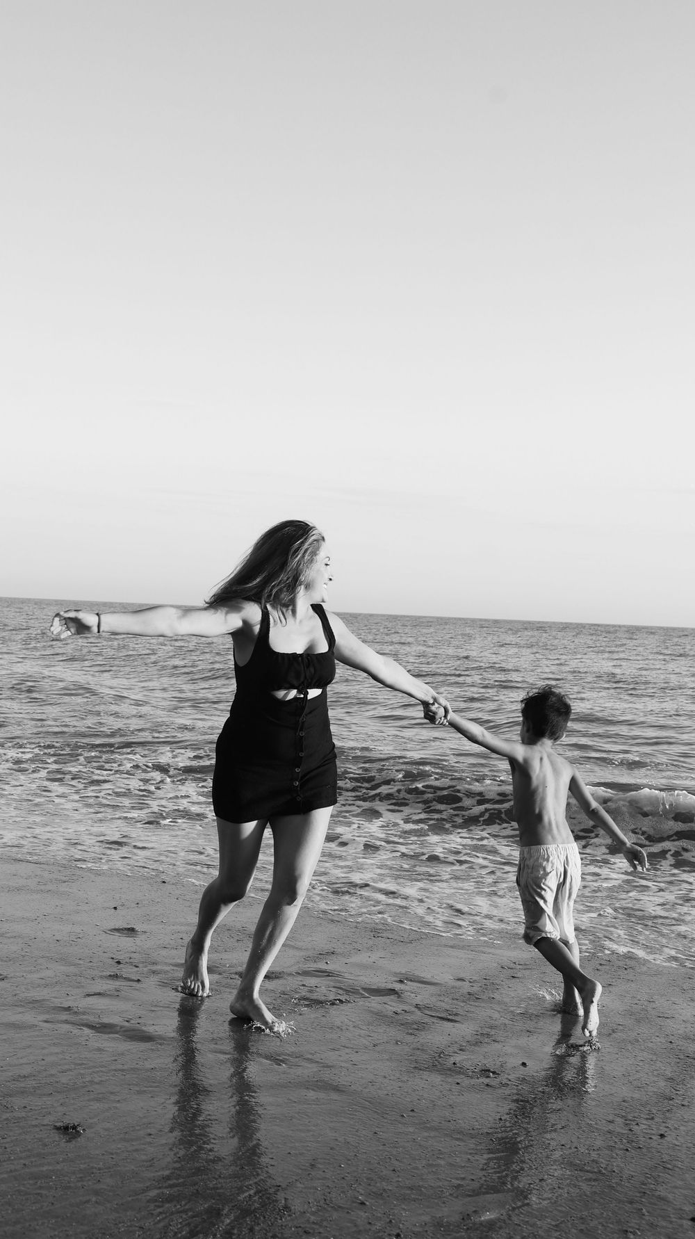 Black and white photo of a woman and child holding hands, playing in ocean waves on a sandy beach.