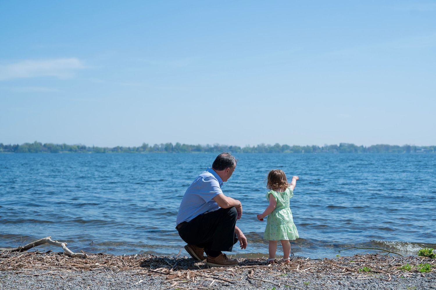 Father crouching with toddler girl on rocky lakeside shore, pointing at calm blue water on sunny day.