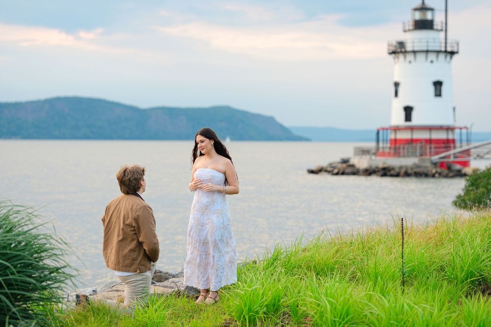 Man kneeling before woman in white dress near lighthouse on Hudson River during romantic marriage proposal.