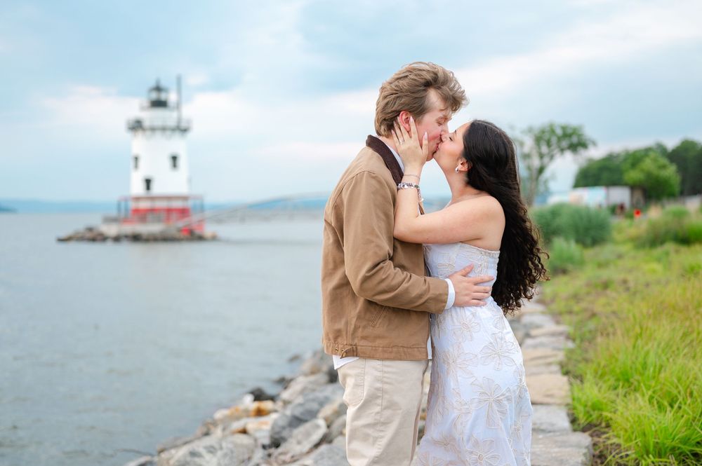 Couple sharing a romantic kiss by a rocky shoreline with a red and white lighthouse in the background.