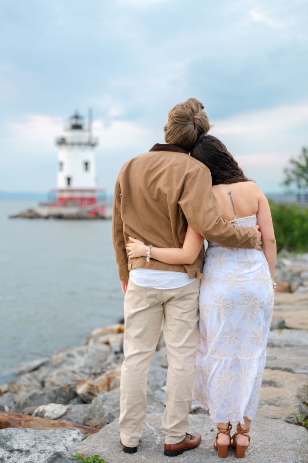 Engaged couple embracing while gazing at lighthouse from rocky waterfront at dusk.