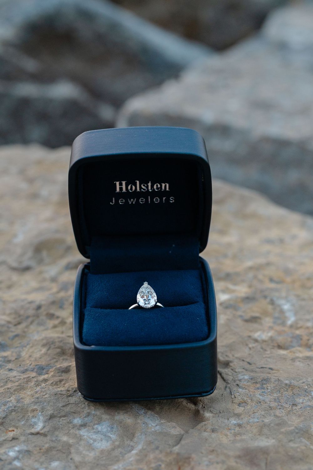 Pear-shaped diamond solitaire engagement ring in black Holsten Jewelers ring box on rocks.
