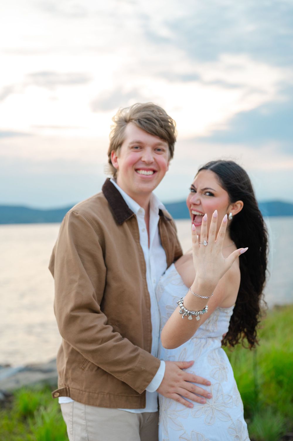 Joyful newly engaged couple showing off diamond ring by the waterfront at sunset.