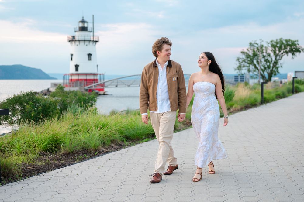 Couple walking hand-in-hand along waterfront path with lighthouse and bridge in background.