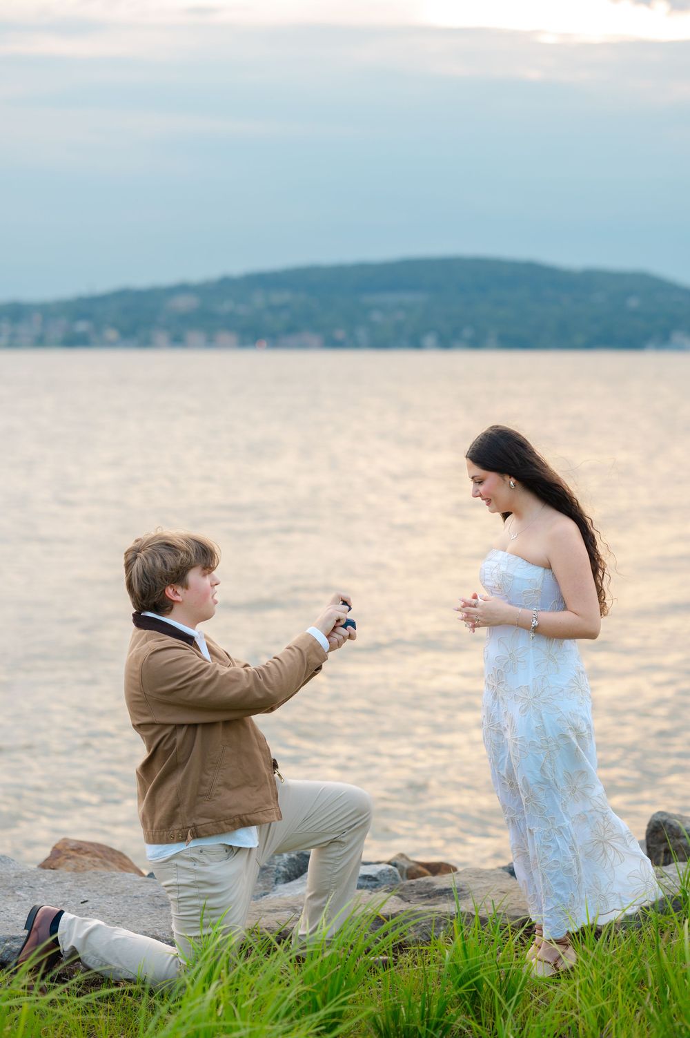 Man on one knee proposing to woman in white dress by waterfront at sunset.