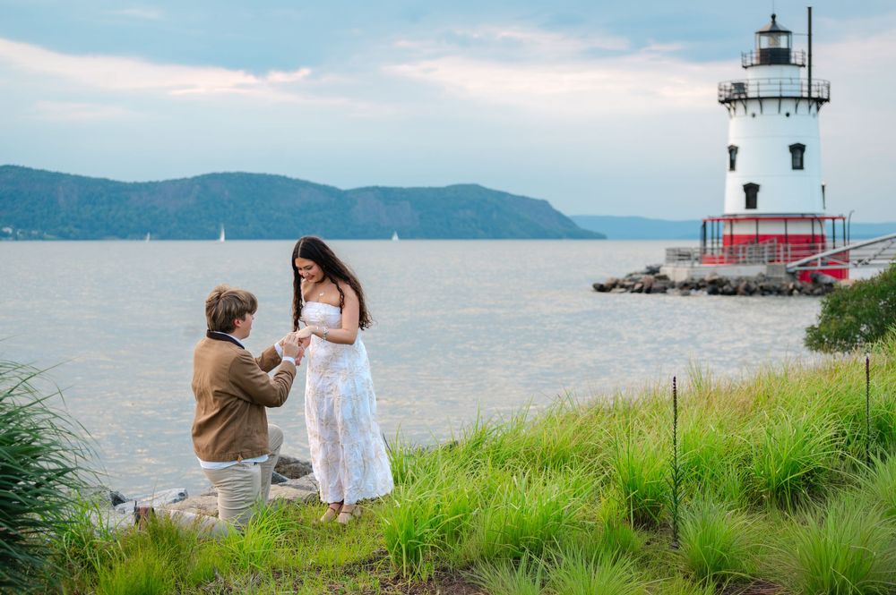 Man proposes to woman in white dress by Hudson River lighthouse on a cloudy day.