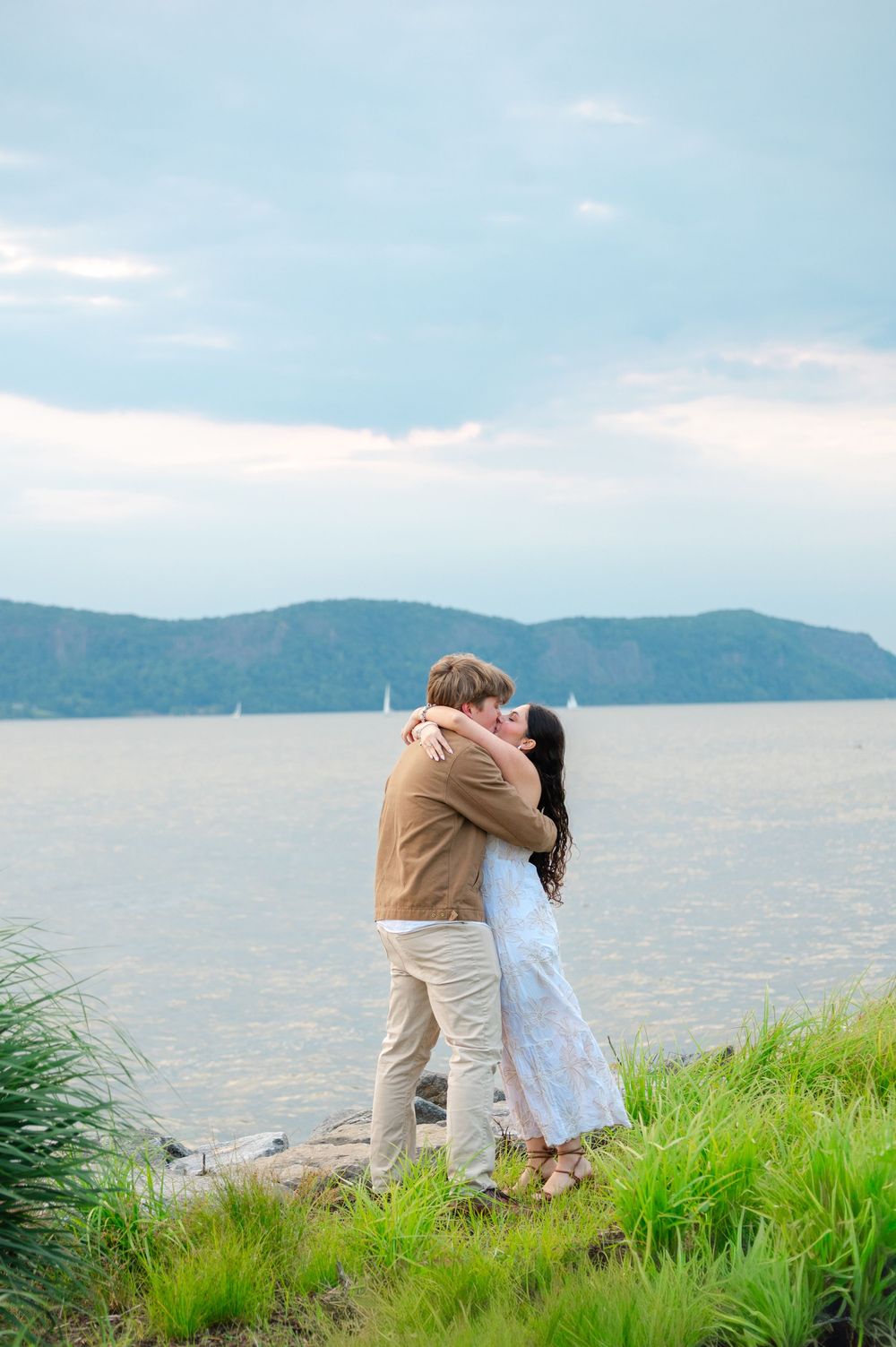 Couple embracing and kissing by a serene lakeside with mountains and sailboats in the background.