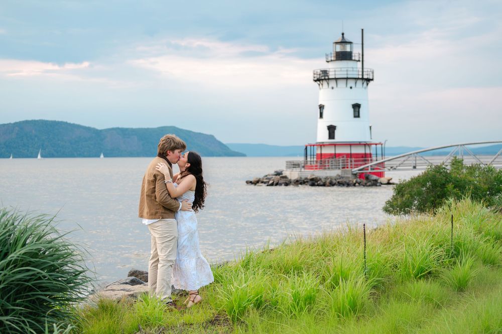 Couple kissing near a white lighthouse by the Hudson River with green grass and mountains in the background.