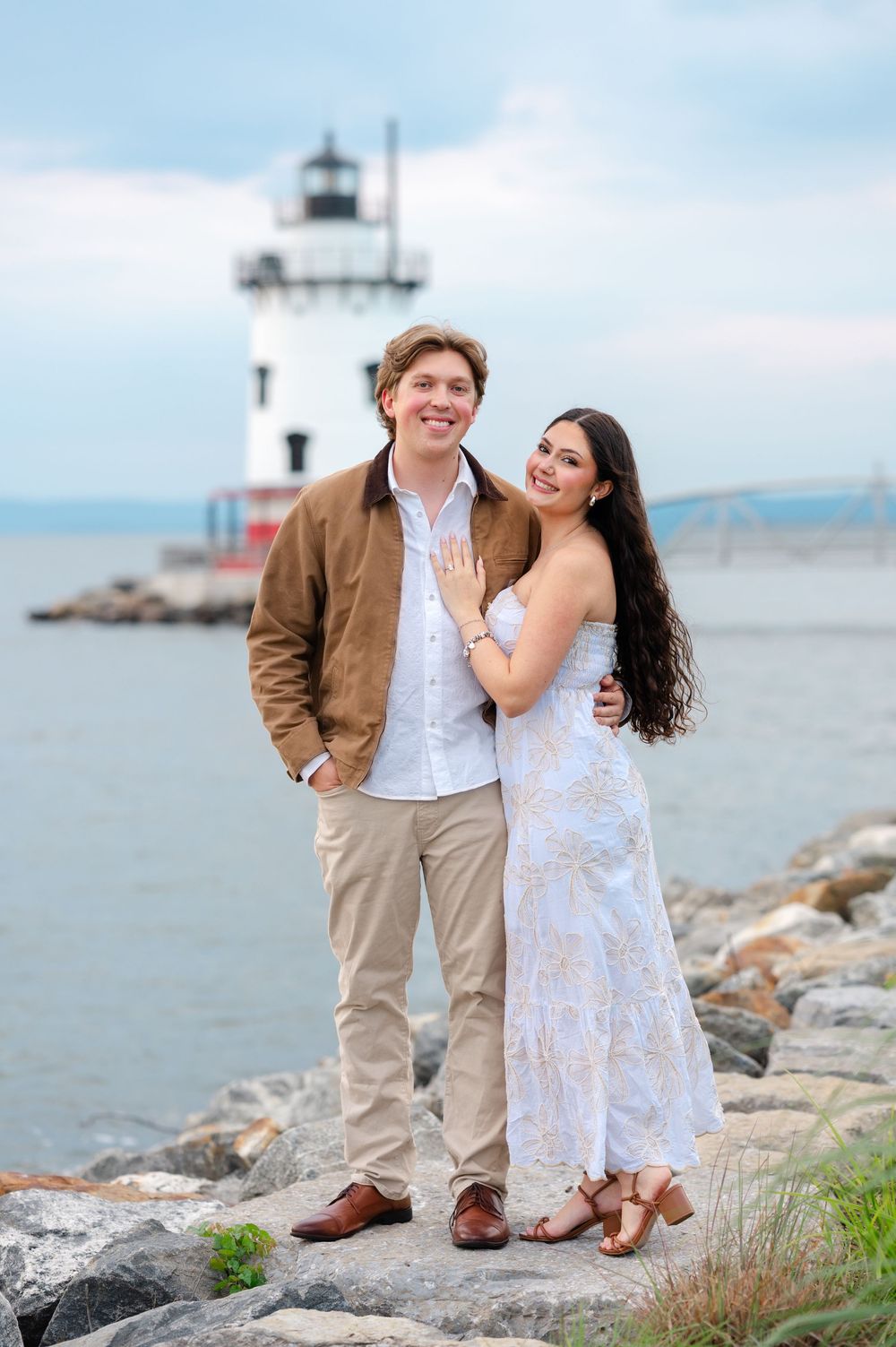 Happy couple posing on rocky shore with lighthouse backdrop during engagement photo session.