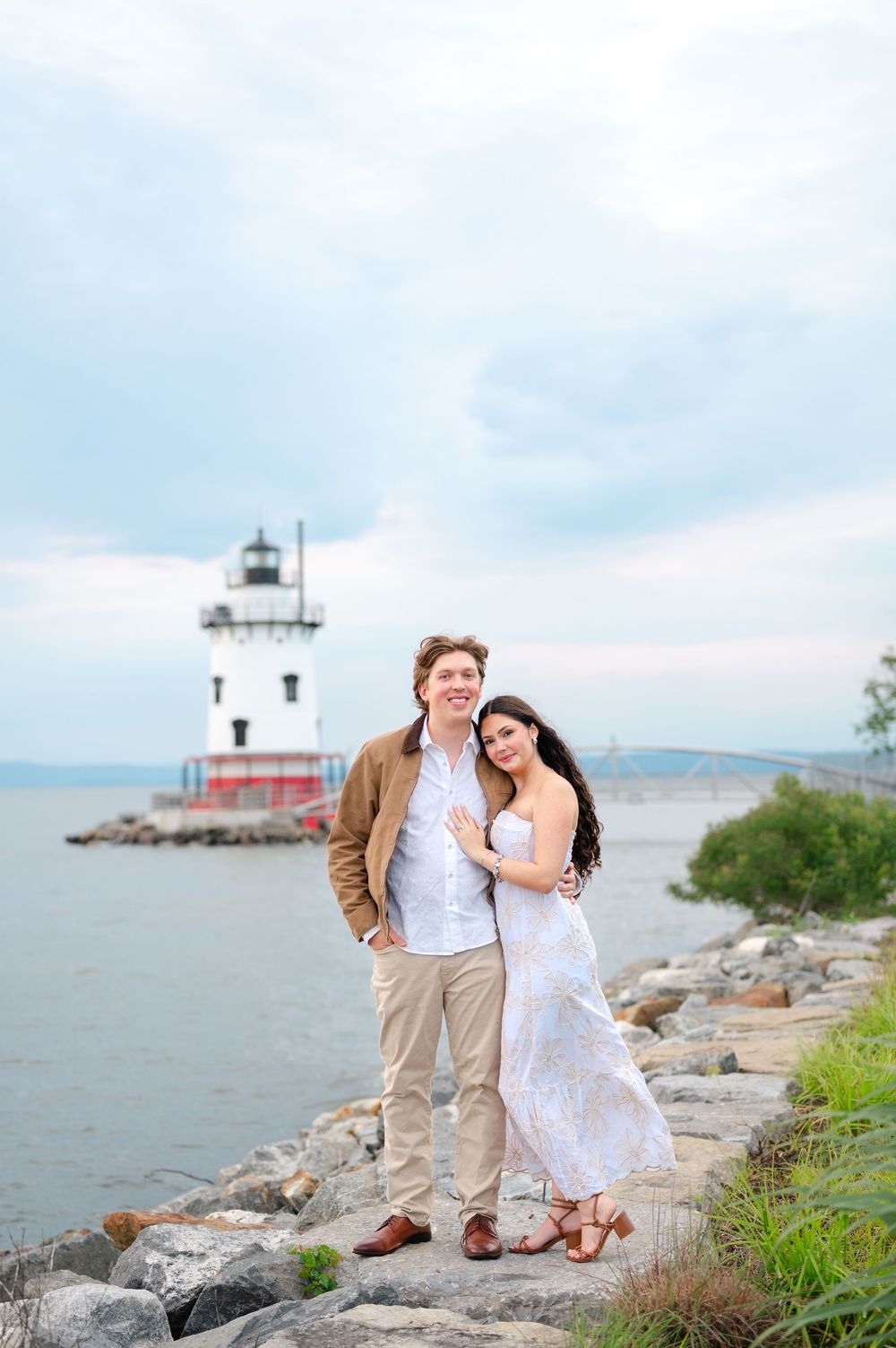Couple poses by rocky shoreline with lighthouse backdrop on cloudy day, woman in white dress, man in brown jacket.