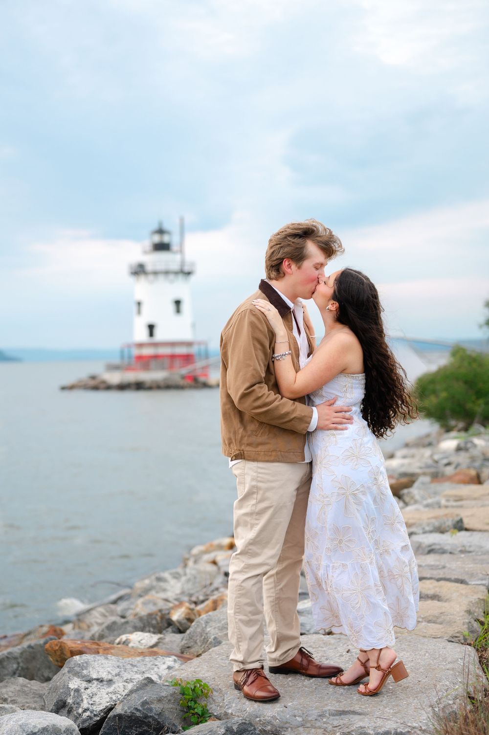 Couple kissing on rocky shoreline with lighthouse in background during engagement photo session.