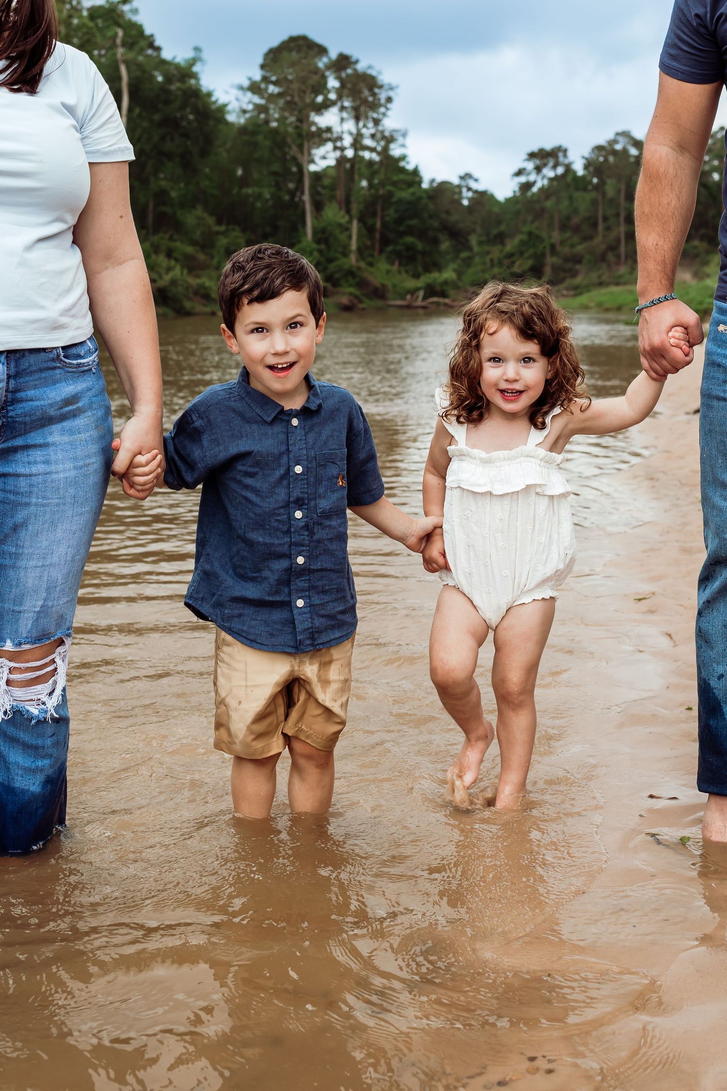 Young siblings holding parents hands while wading in shallow river.