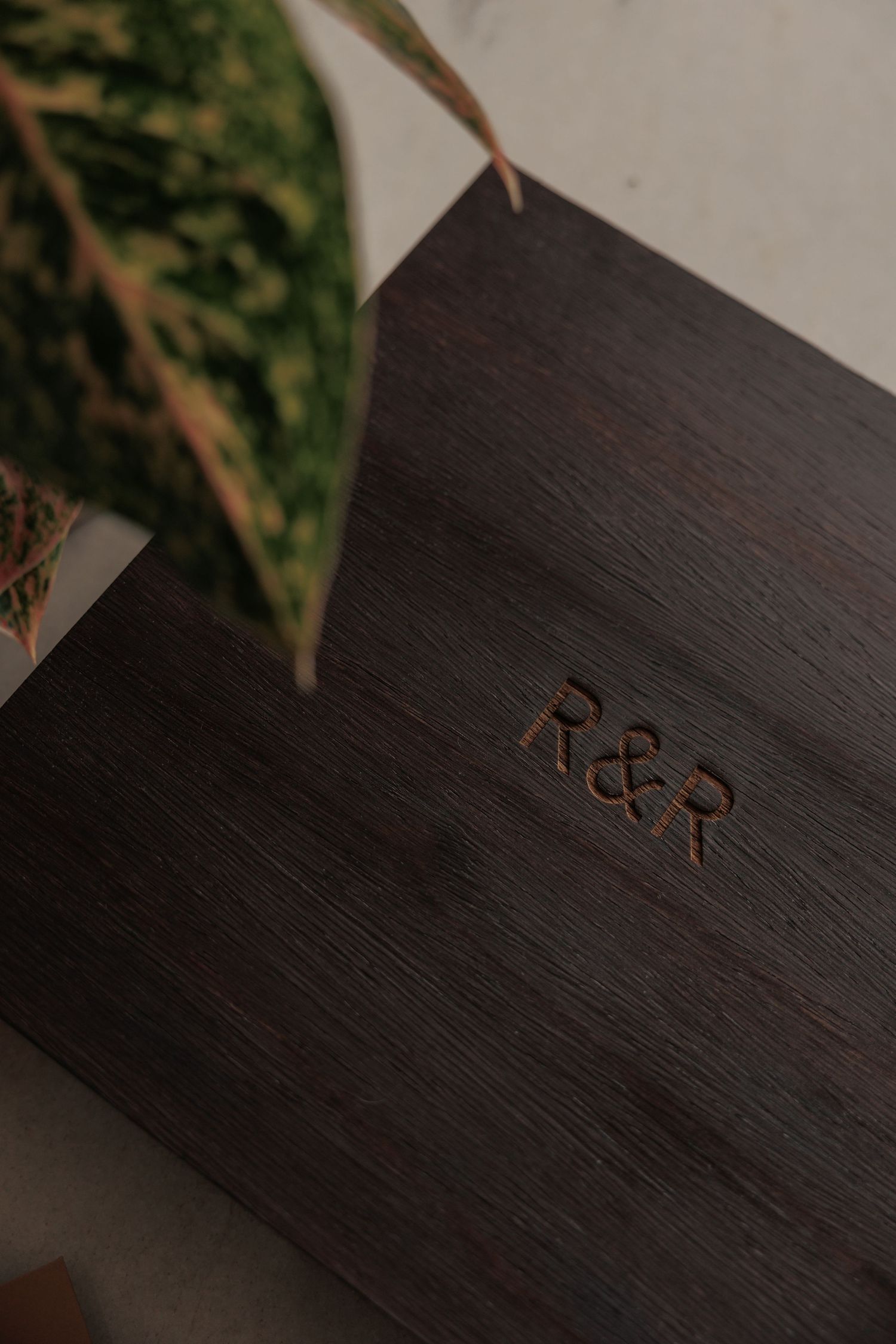 Dark wooden box with engraved R&R monogram initials, surrounded by green tropical leaves.