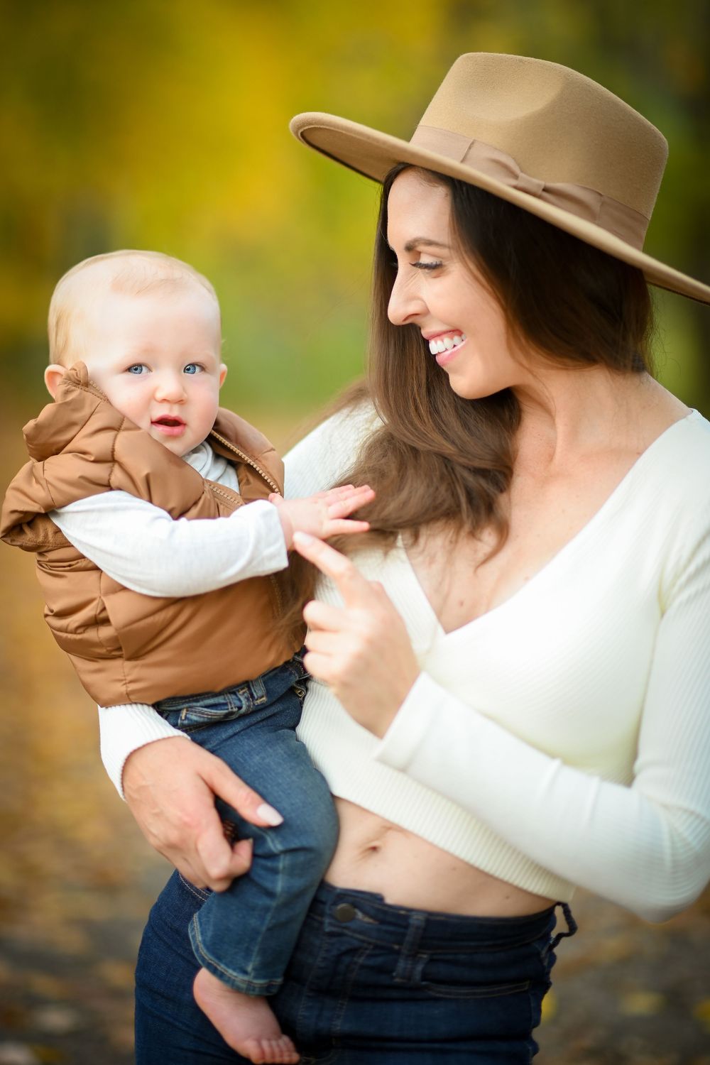 A smiling woman in a tan hat and white crop top holds an adorable baby in a brown vest outdoors in autumn.