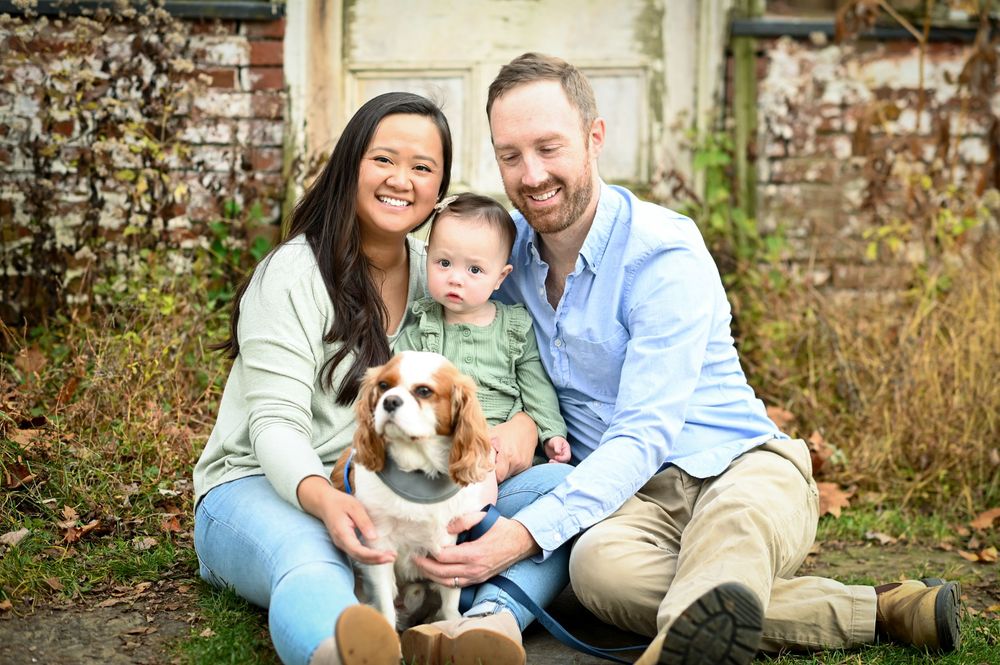A smiling family with a baby and Cavalier King Charles Spaniel dog poses outdoors in a lush garden setting.