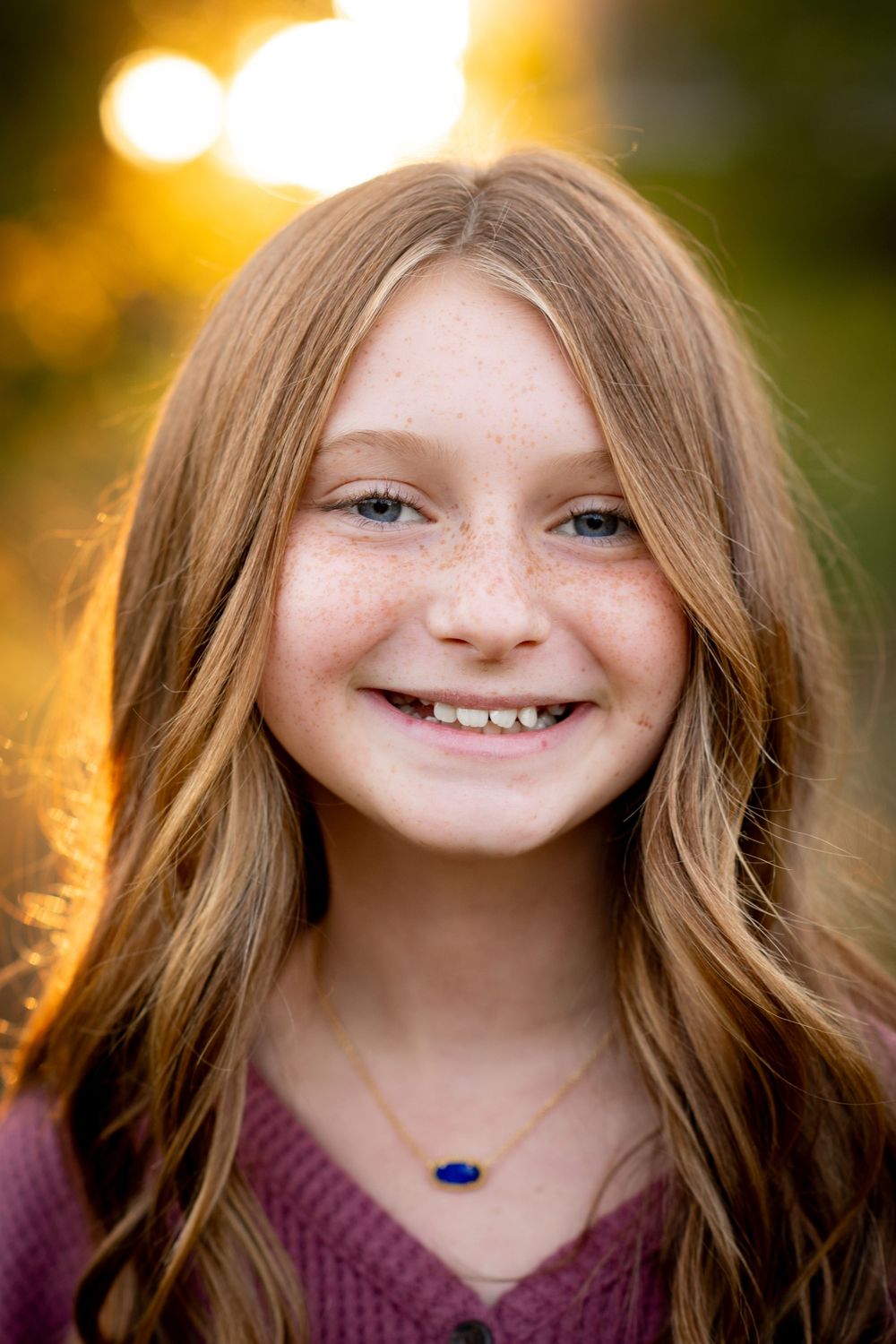 Smiling young girl with freckles and long auburn hair wearing a blue pendant necklace in golden hour light.