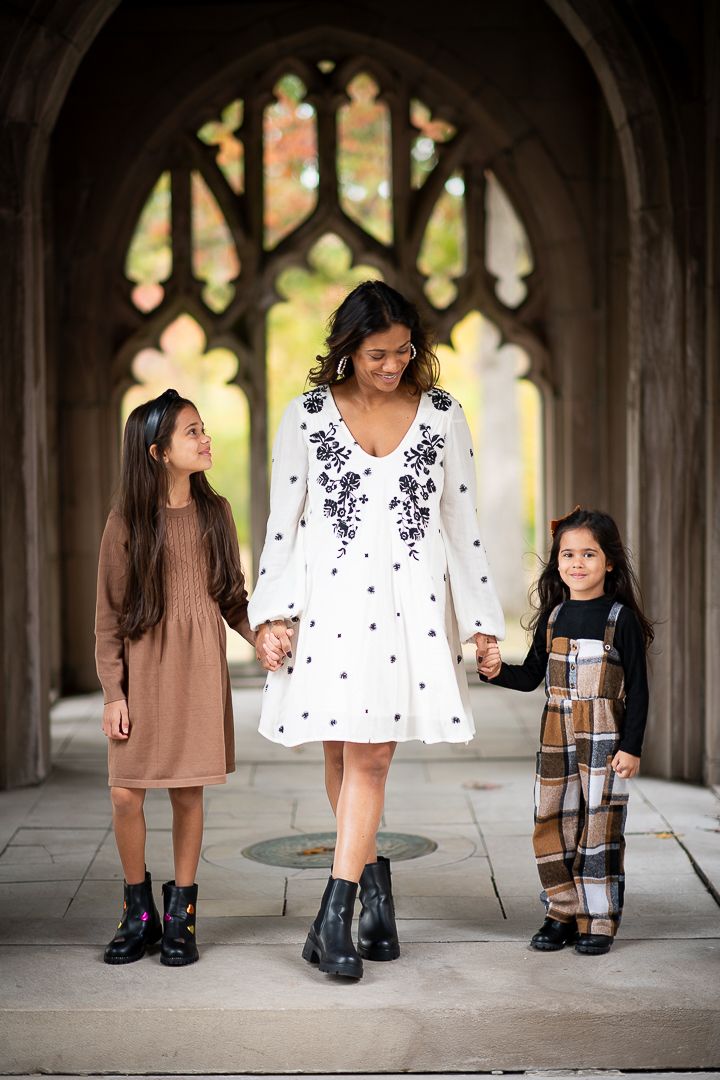 A woman in a white embroidered dress holds hands with two young girls in a gothic archway setting.