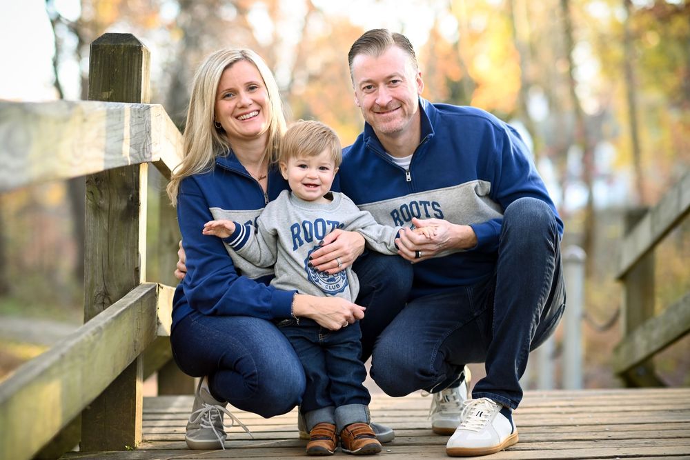 Family of three smiling on a wooden bridge in autumn, wearing matching Roots sweaters and jeans.