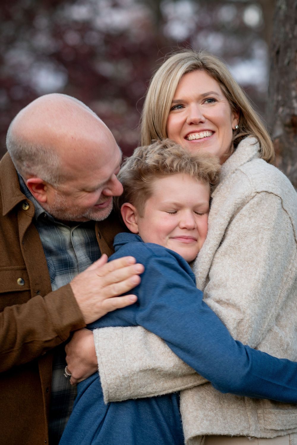 A happy family of three shares a warm embrace outdoors, with parents hugging their young son in winter attire.