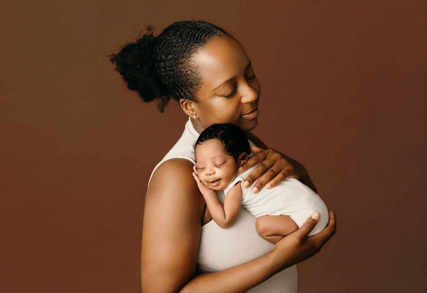 A Black mother tenderly holds her sleeping newborn baby against her chest in a warm brown studio portrait.