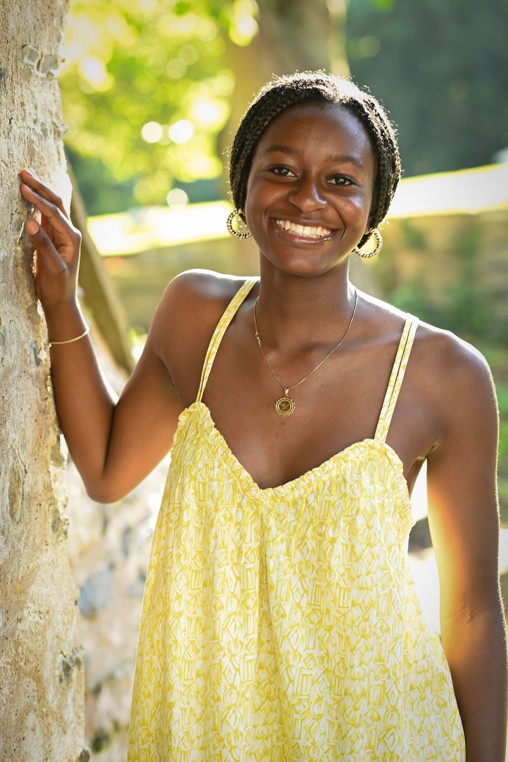 Smiling young Black woman in yellow sundress leaning against tree outdoors in sunlight.