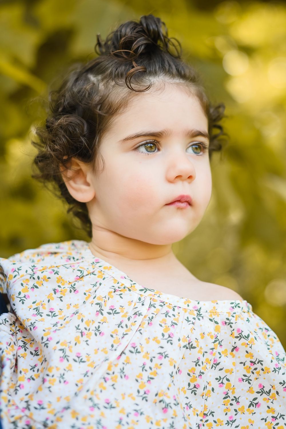 Toddler girl with curly dark hair and green eyes wearing a floral dress, gazing thoughtfully outdoors.