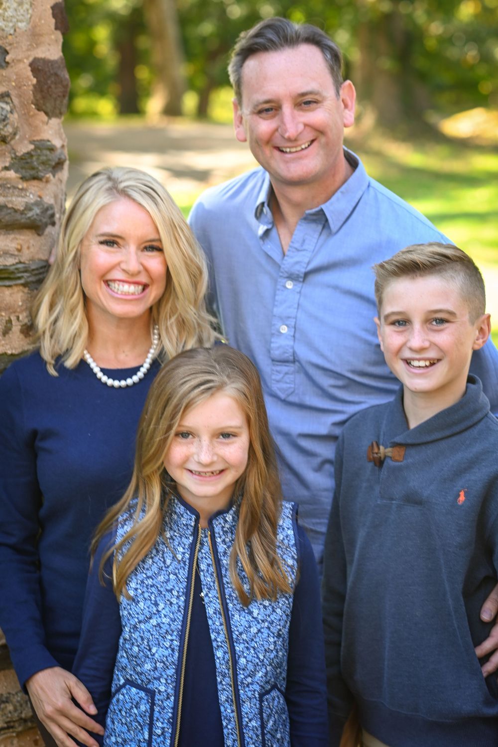 Happy family of four posing outdoors near a stone wall, parents with two children smiling in coordinated blue outfits.