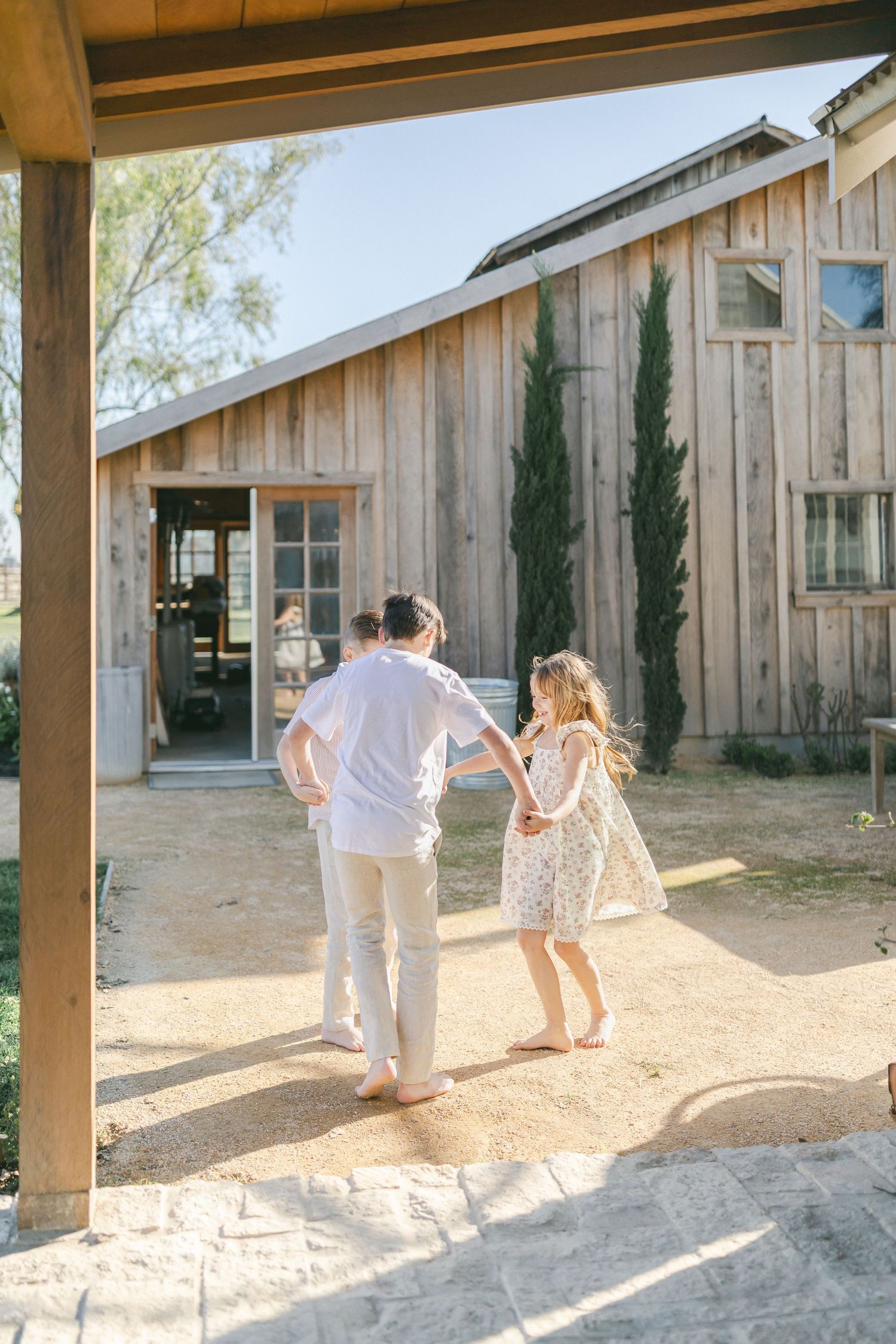 A family of five poses barefoot in front of a rustic wood building, smiling warmly in soft pastel outfits.