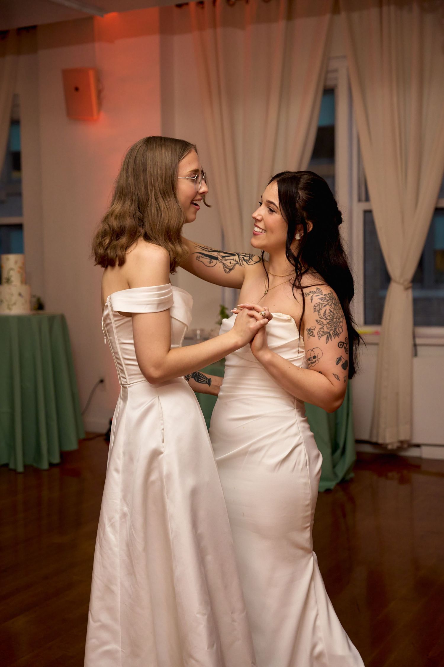 Two brides in white satin gowns share their first dance, smiling at each other during wedding reception.