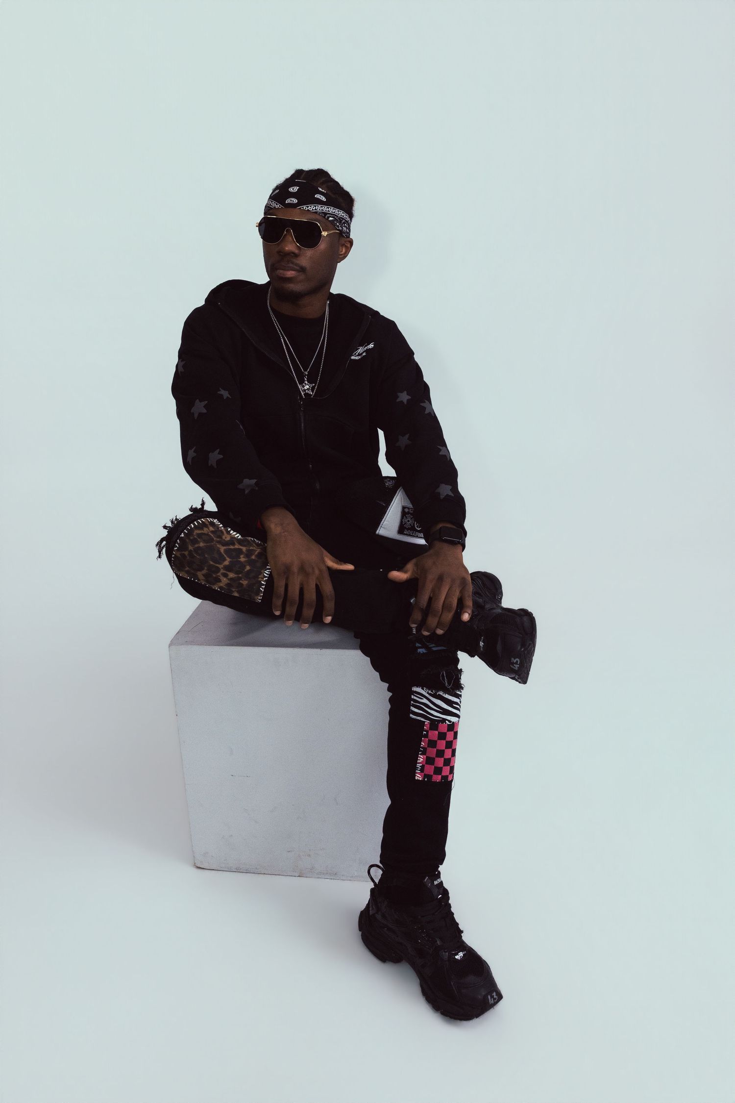 Stylish man in black outfit with checkered pants sits on concrete cube against light background.