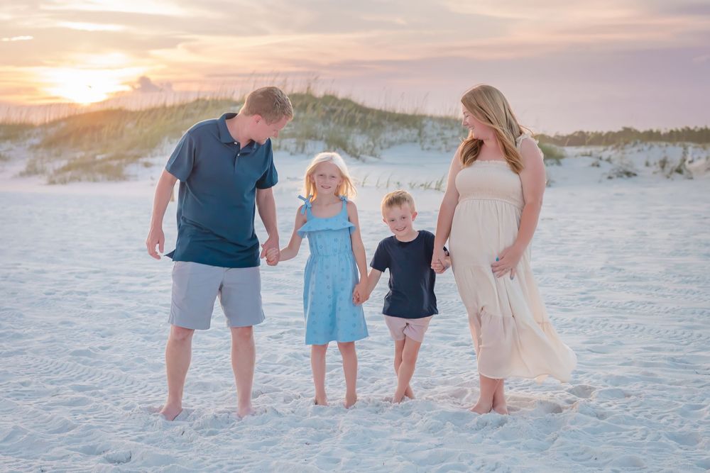 Happy family of four holds hands on white sand beach during colorful sunset.