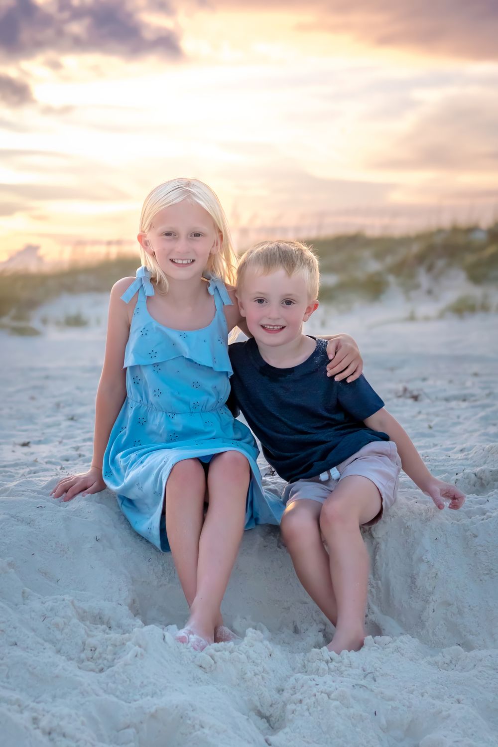 Two blonde siblings smile and hug while sitting on sandy beach at sunset.