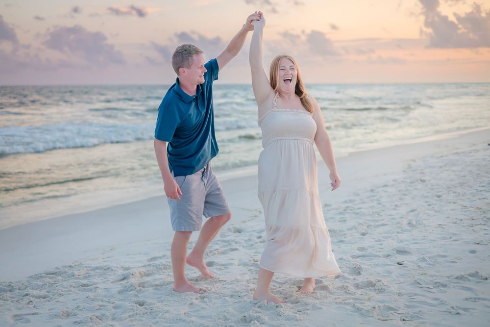 Couple dancing joyfully on a sandy beach at sunset, man spinning woman in white dress by the ocean.